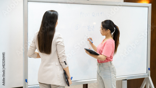 Two women are standing in front of a white board, one of them writing on it