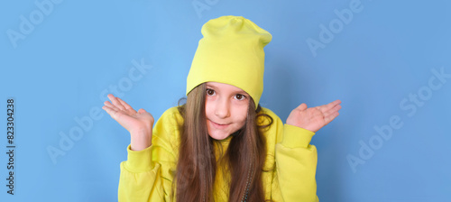 Doubtful girl, refuses, shrugs, isolated blue color background. I don't know. Confused, questioning child hesitantly raises his hands.