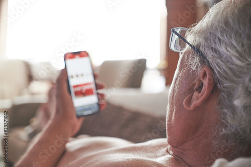 Retired man without a shirt reads the news from a digital newspaper through the screen of his mobile phone comfortably on the sofa at home.