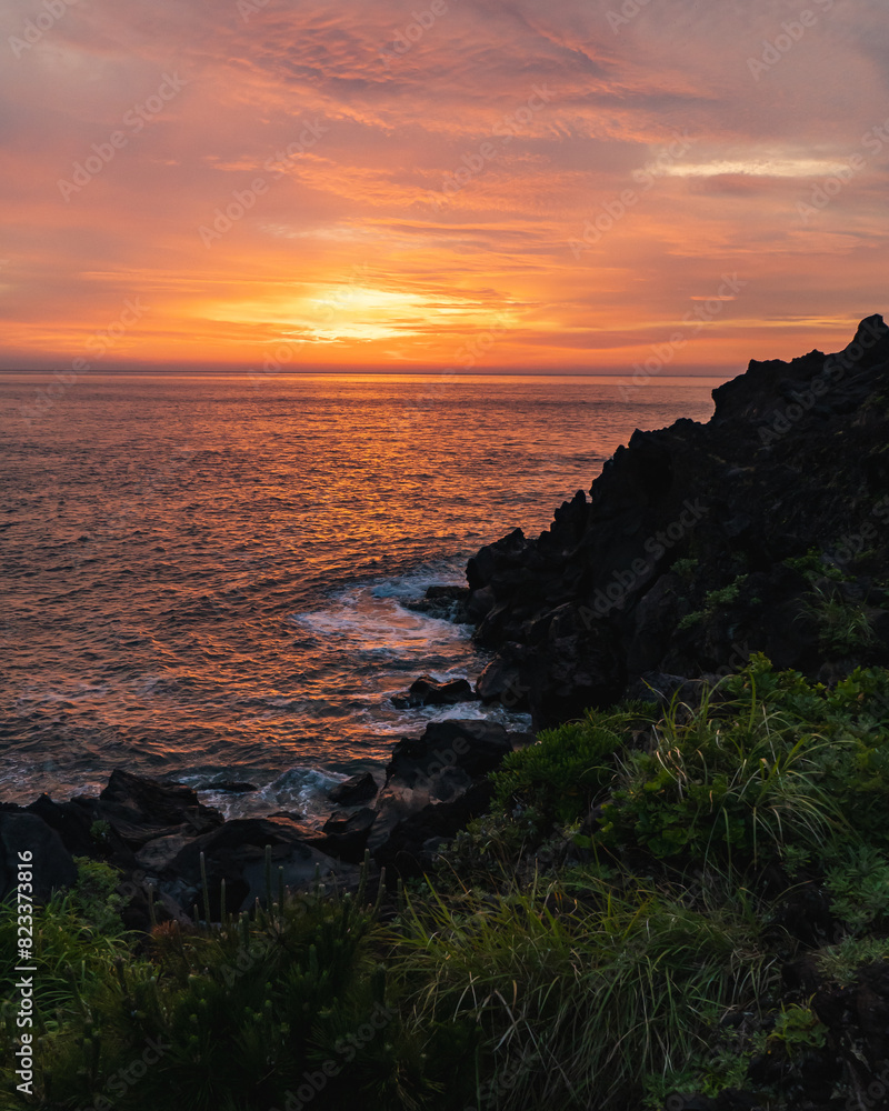 A beautiful coastal area in eastern part of Japan. A clear blue sky and an orange sunrise reflects on the sea water. A big rock formation in the coast line. Island from the distance. A beautiful coastal area in eastern part of Japan. A clear blue sky and an orange sunrise reflects on the sea water. A big rock formation in the coast line. Island from the distance.