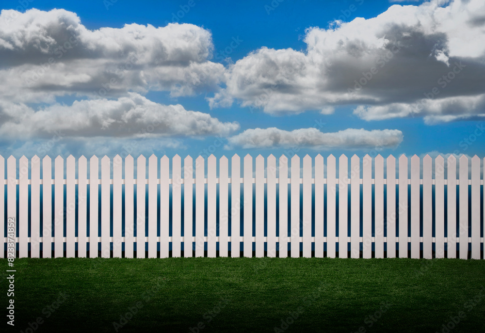 White picket fence with green grass and fluffy clouds in the background White picket fence with green grass and fluffy clouds in the background