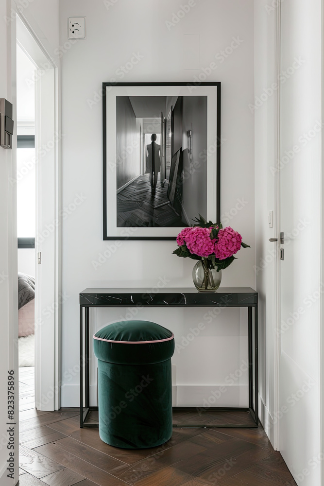 Fototapeta premium A small black console table with an emerald velvet stool is located in the center of an empty white wall. On it, there is a photo frame and pink peonies. The photo depicts a modern apartment interior