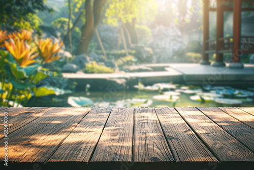 Wooden deck with shadows facing a Japanese garden pond surrounded by green plants blooming flowers and a small pavilion under soft sunlight photo