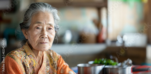 Elderly woman in a kitchen, traditional attire, thoughtful expression, soft background, day, kitchen items visible
