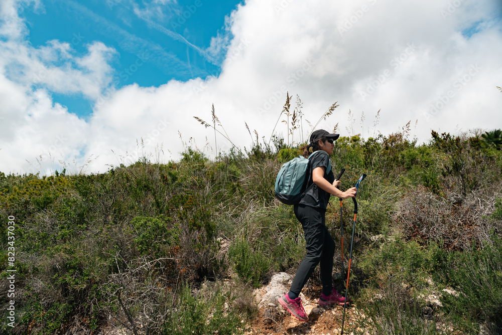 Obraz premium Woman hiker walking on a scenic mountain trail under a blue sky with clouds.