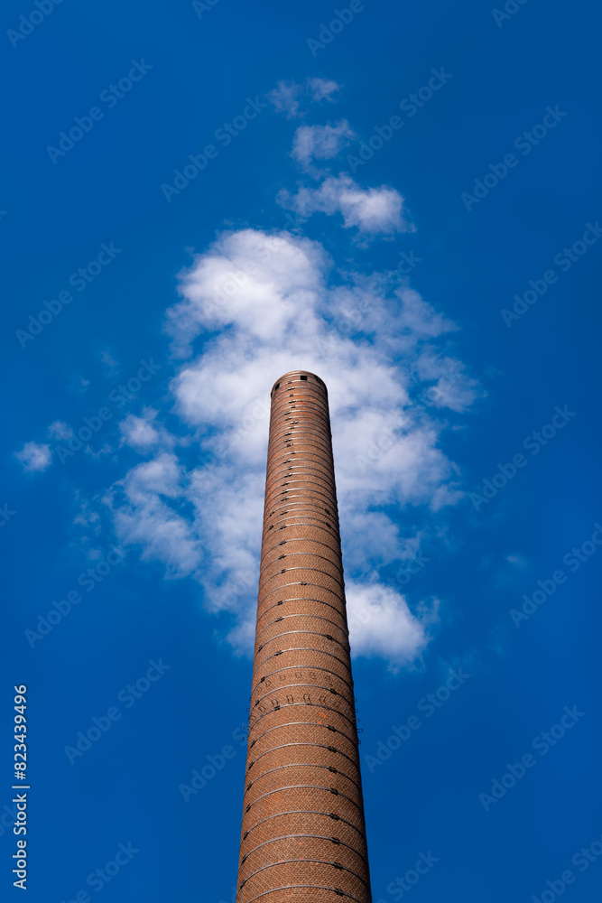 Smoke stack of an old factory with iron steps leading upwards and red bricks on a sunny blue sky day. A white fair-weather cloud directly behind the chimney looks like a cloud of exhaust fumes.