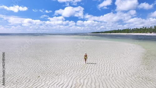 Aerial view of walking young woman on the sandbank in ocean, white sand, blue sea during low tide at sunny summer day in Zanzibar island. Top view of girl, sand spit, bay, clear water, sky with clouds