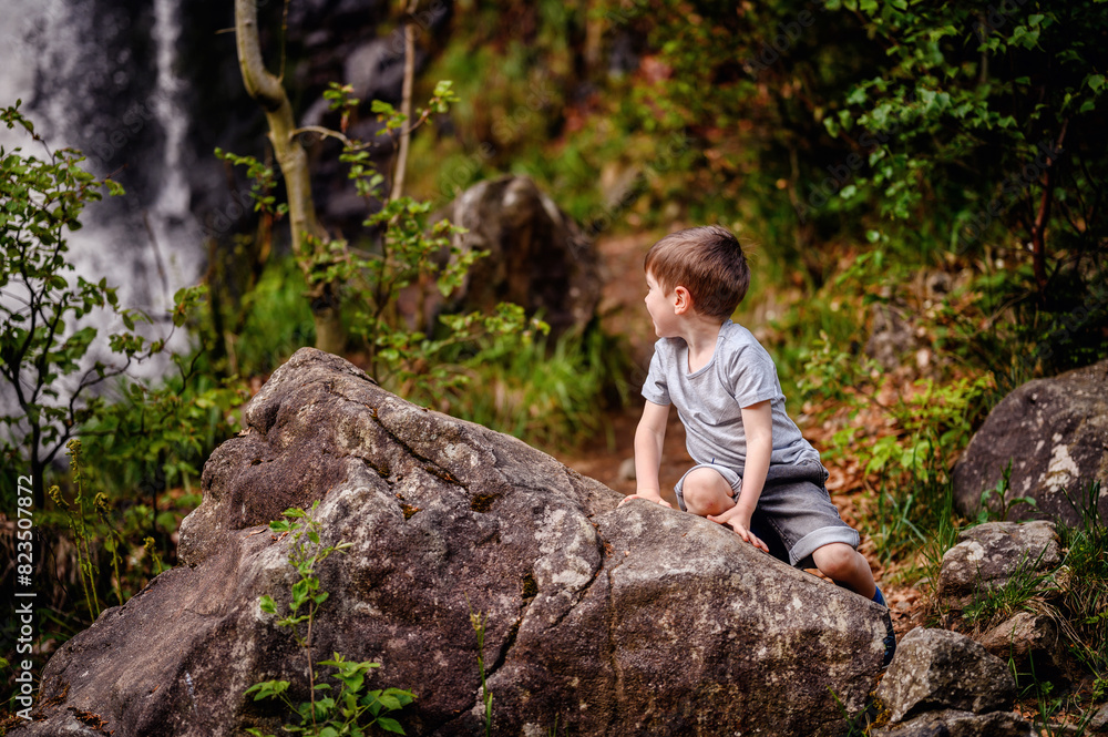 Smiling young boy in a gray t-shirt and denim shorts climbing large rocks in a forested area. Capturing a moment of joyful adventure and exploration in nature. Smiling young boy in a gray t-shirt and denim shorts climbing large rocks in a forested area. Capturing a moment of joyful adventure and exploration in nature.