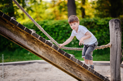 A young boy in a white shirt and denim shorts carefully climbs a wooden ramp using a rope at a playground, enjoying outdoor playtime surrounded by greenery.