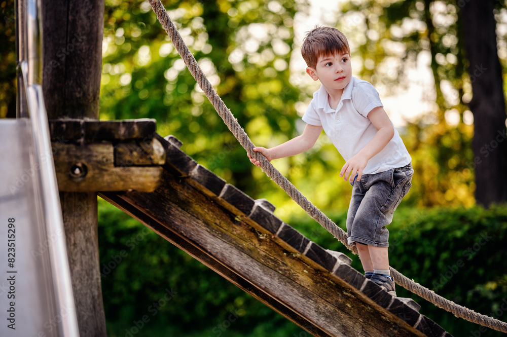 A young boy in a white shirt and denim shorts enthusiastically climbs a wooden ramp using a rope at a playground, set against a background of lush greenery on a sunny day. A young boy in a white shirt and denim shorts enthusiastically climbs a wooden ramp using a rope at a playground, set against a background of lush greenery on a sunny day.