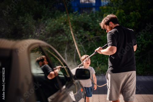 A father teaching his young son to wash a car with a high-pressure hose at a self-service station. The boy looks up at his dad with admiration, sharing a bonding moment © Иванна Емельянова