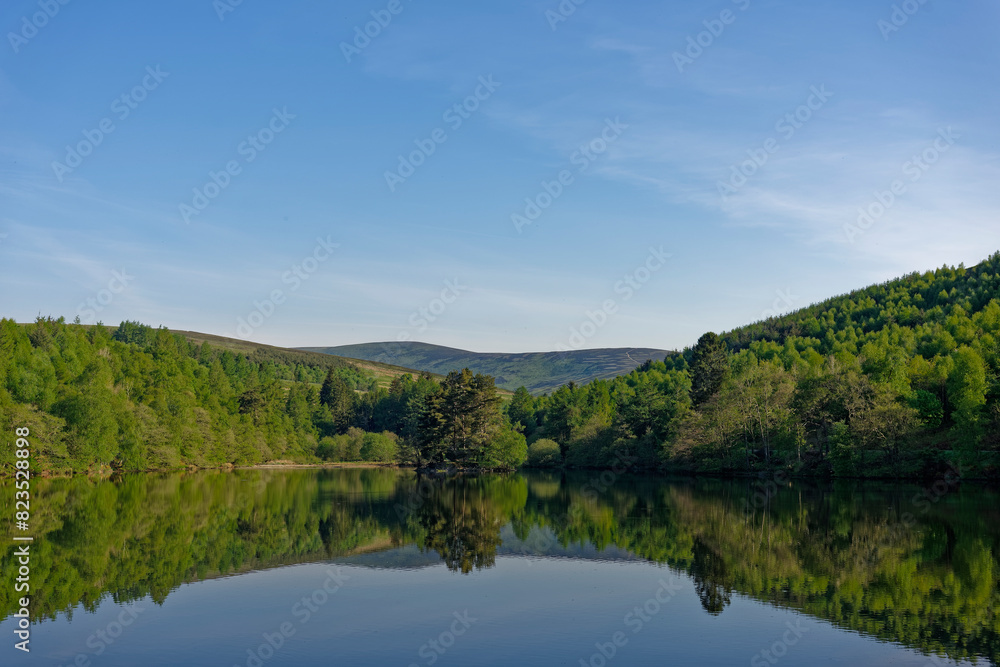 A tranquil view of the Glen Ogil Reservoir with the hills of the Angus Glens in the background, and Reflections of the Wooded margins in the calm water.