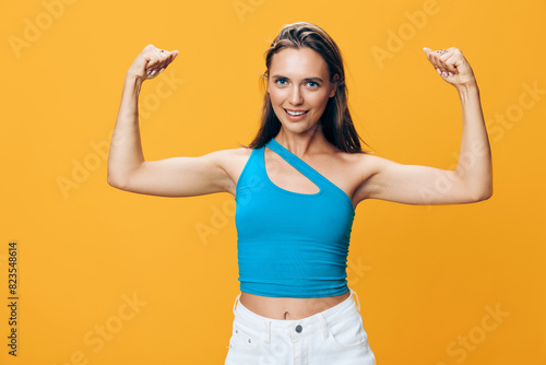 Powerful woman showing strength and confidence in blue top flexing biceps on yellow background
