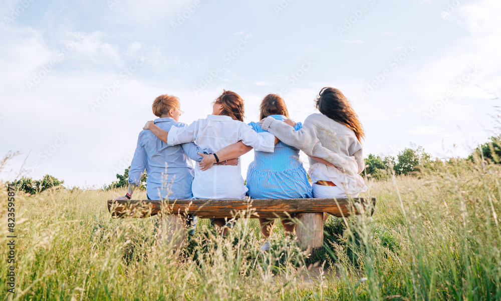Four smiling women dressed light summer clothes harm embracing sitting on the meadow bench during outdoor walking. Woman's friendship, relations, and happiness concept image. Four smiling women dressed light summer clothes harm embracing sitting on the meadow bench during outdoor walking. Woman's friendship, relations, and happiness concept image.