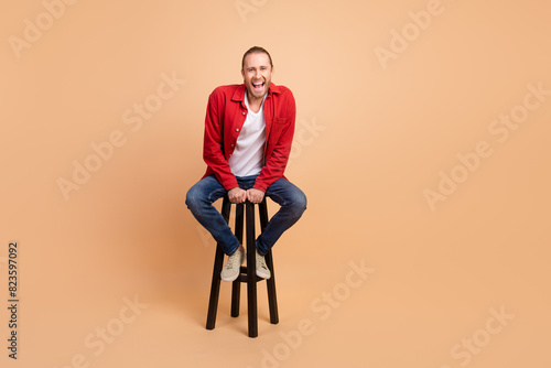 Full size photo of nice young man sit stool laugh wear red shirt isolated on beige color background