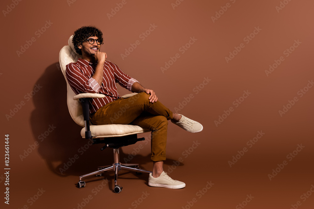 Full body portrait of nice young man sit chair look empty space wear striped shirt isolated on brown color background