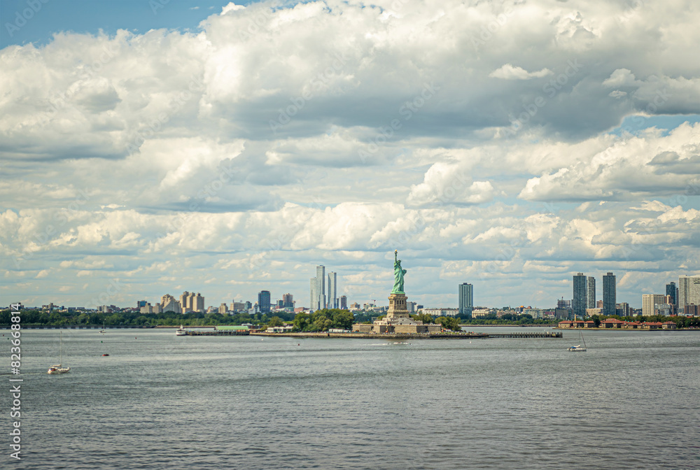 Naklejka premium New York, NY, USA - August 1, 2023: Statue of Liberty on its island in wide landscape showing New Jersey coastline under blue cloudscape