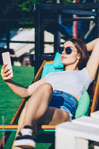Charming young woman with long hair and sunglasses making selfie photo on front camera of modern smartphone lying on sunbed.Hipster girl resting and sunbathing while publishing new posts in blog © BullRun