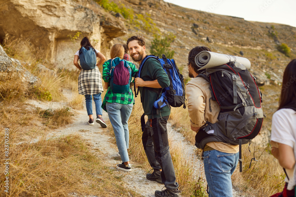 Full length portrait of a smiling hiker surrounded by a group of tourists friends. The traveler stands against the backdrop of mountain, during group hike at holiday vacation.