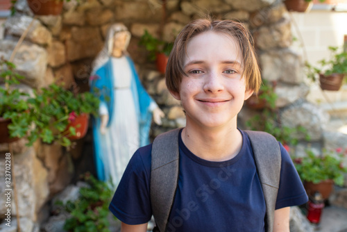 An 11-year-old smiling Catholic boy with a backpack against the background of a statue of the Virgin Mary. photo