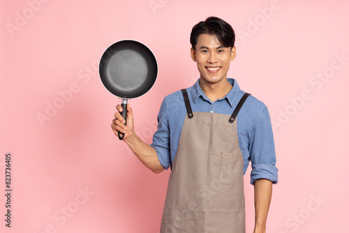 Young Asian man chef wearing kitchen apron cooking and holding pan and spatula isolated on pink background photo