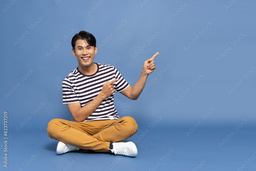 Young Asian man smiling and pointing to empty copy space and sitting on floor isolated on blue background Young Asian man smiling and pointing to empty copy space and sitting on floor isolated on blue background