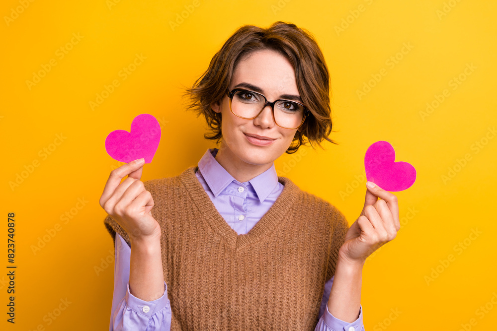 Portrait of lovely adorable woman wear knit waistcoat in glasses hands hold two pink heart postcards isolated on yellow color background Portrait of lovely adorable woman wear knit waistcoat in glasses hands hold two pink heart postcards isolated on yellow color background