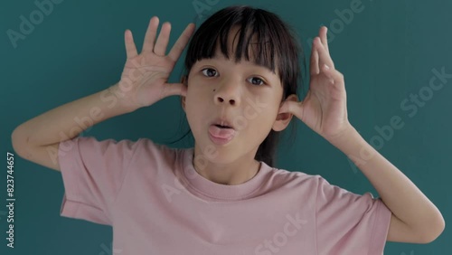 Portrait of a cute and healthy-looking Thai Asian kid girl, aged 8 to 10 years, happily holding her hands behind her ears and sticking out her tongue and eyes. background is green screen.