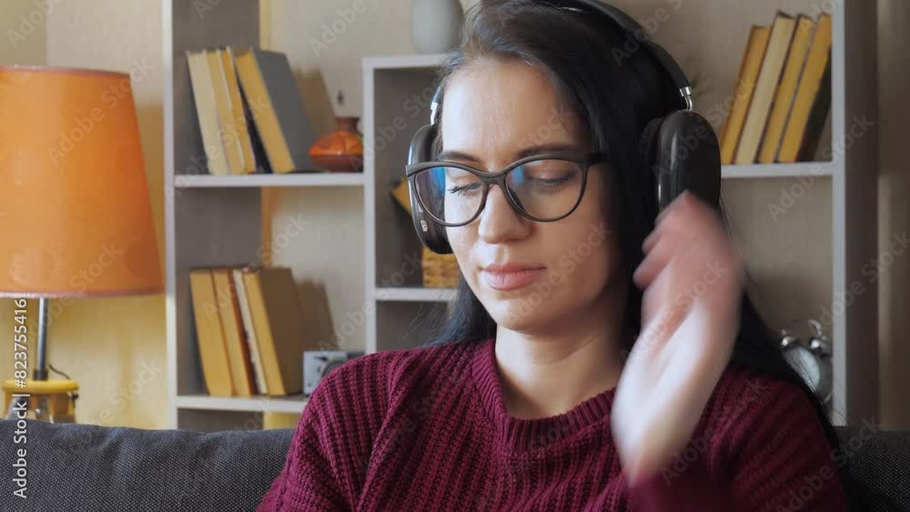 A beautiful young girl with glasses, with black straight hair, in a burgundy blouse, puts on headphones, looks for her favorite melody, starts listening, emotionally swaying to the beat. A beautiful young girl with glasses, with black straight hair, in a burgundy blouse, puts on headphones, looks for her favorite melody, starts listening, emotionally swaying to the beat.
