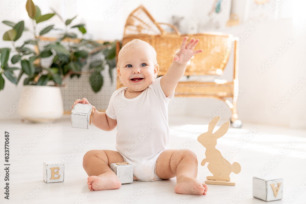 Fototapeta premium a little baby girl of six months with blue eyes in a white bodysuit plays on the floor of the children's room with wooden toys cubes smiles and rejoices, early development of the child