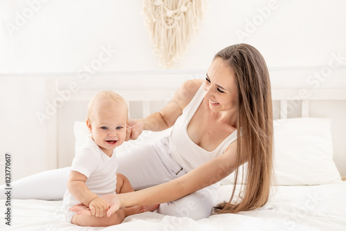 a young beautiful mother with a baby hugs and kisses him or plays with him, having fun on the bed in a bright bedroom, happy motherhood with maternity leave, maternal love and care