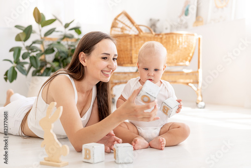 mom and baby play with wooden toys cubes having fun together in the children's bright room, happy motherhood with maternity leave, mom teaches the baby, early development of the child