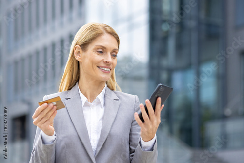 A confident businesswoman in a grey suit holds a credit card and smartphone while standing in a modern urban business district. She appears to be engaging in a financial or online transaction.