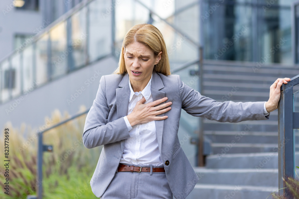 A blonde woman, in a formal grey suit, clutches her chest in pain while standing outside a modern office building, appearing to be experiencing a heart attack or severe chest pain.