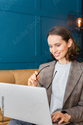 Beautiful woman with brown hair sitting on the sofa, smiling and working on a laptop.