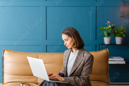 Beautiful woman with brown hair sitting on the sofa and working on a laptop.