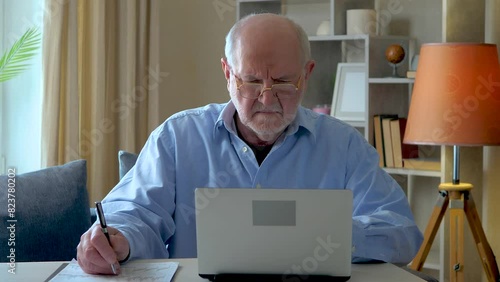 An elderly businessman, gray-haired in a blue shirt, at his desk, working on a computer, reconciling the data of the accounting report, writing it down in a document. Slow motion. photo