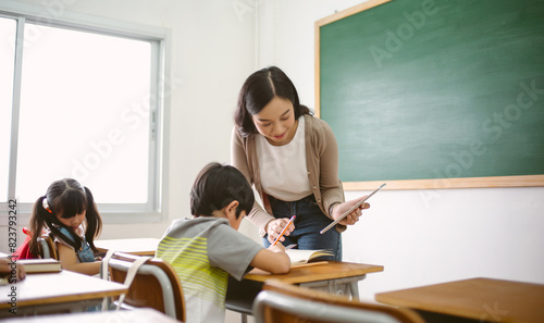 Happy Asian female teacher helping schoolboy with digital tablet in computer in IT Class. Education, elementary school, learning and technology concept.