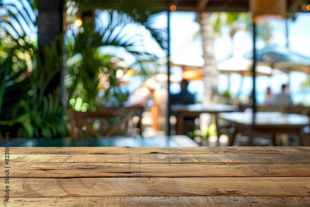 A wooden table in the foreground with a blurred background of a beachside cafe. The background shows sandy beach views through large windows, casual seating, tropical plants, and customers A wooden table in the foreground with a blurred background of a beachside cafe. The background shows sandy beach views through large windows, casual seating, tropical plants, and customers