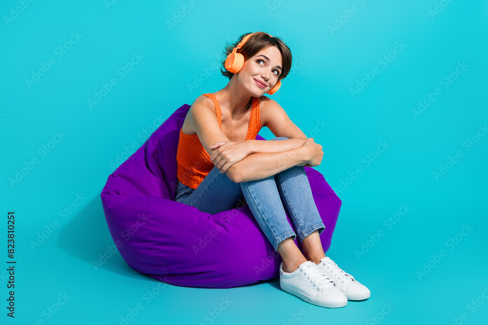 Full size photo of gorgeous good mood girl sit on bean bag in headphones look at promo empty space isolated on blue color background Full size photo of gorgeous good mood girl sit on bean bag in headphones look at promo empty space isolated on blue color background