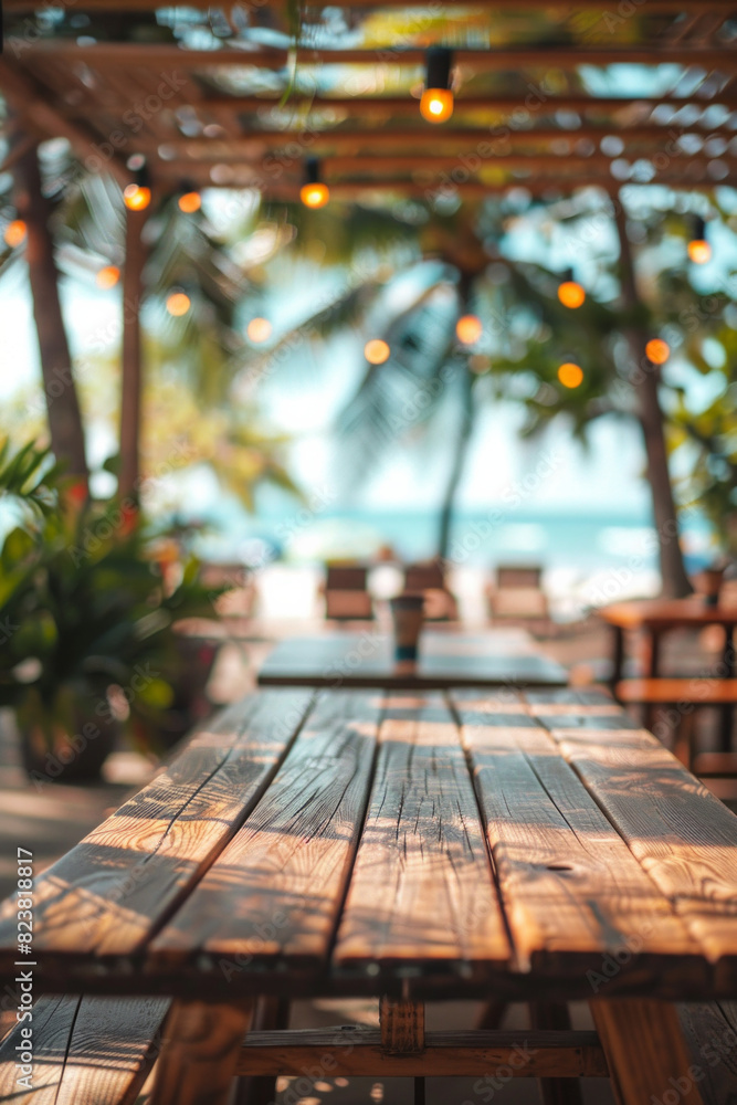 A wooden table in the foreground with a blurred background of a beachside cafe. The background shows sandy beach views through large windows, casual seating, tropical plants, and customers A wooden table in the foreground with a blurred background of a beachside cafe. The background shows sandy beach views through large windows, casual seating, tropical plants, and customers