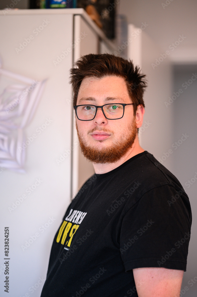 Obraz premium Portrait of a man with glasses and a beard standing indoors, looking at the camera with a neutral expression.