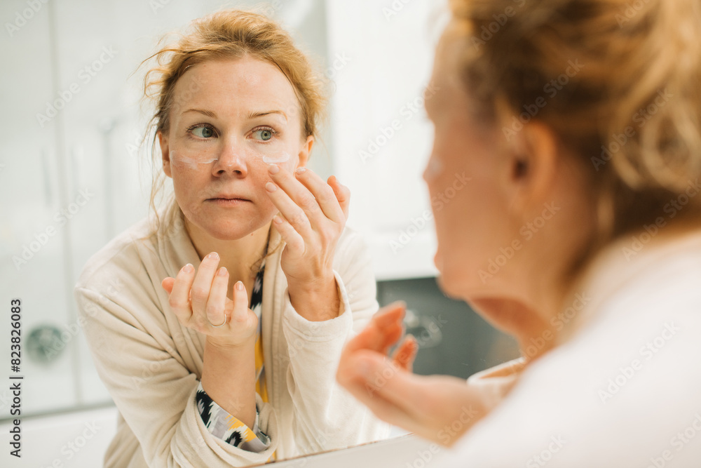 Woman applying moisturazer on her face in front of a mirror Woman applying moisturazer on her face in front of a mirror