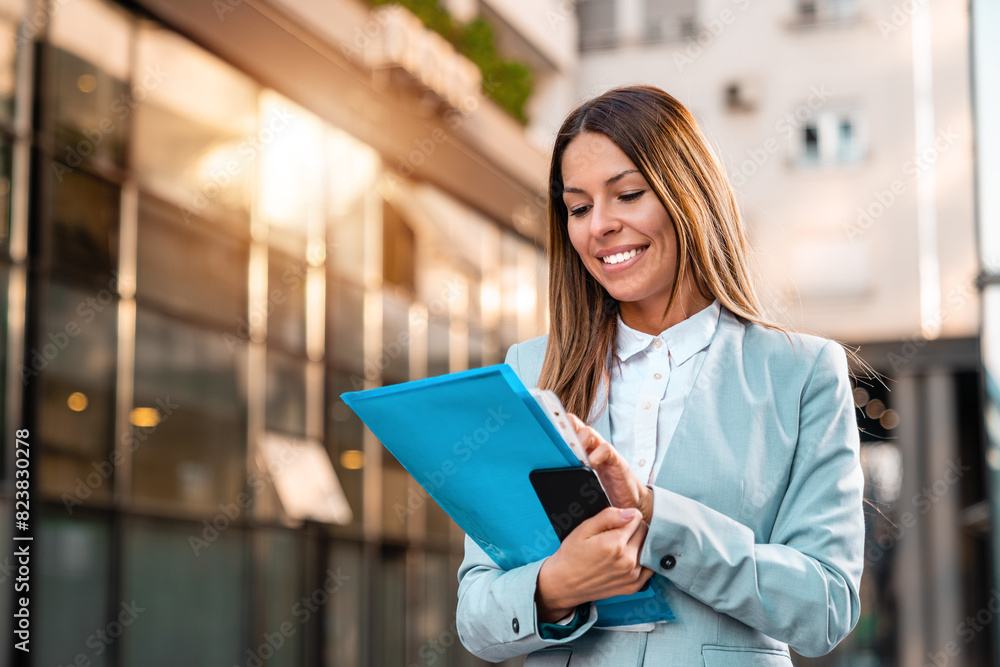 A smiling female office worker with a folder with files and a mobile phone in her hands is walking on the way to work. A smiling female office worker with a folder with files and a mobile phone in her hands is walking on the way to work.