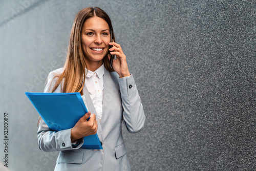 Female real estate agent talking on phone while walking with file folder outdoors. photo