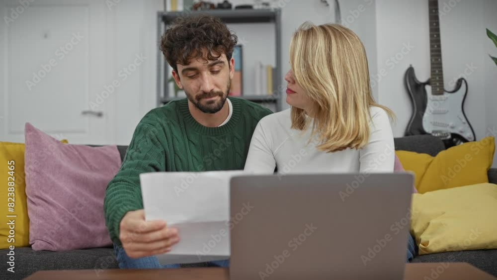 A man and woman examine documents beside a laptop in a cozy living room, reflecting modern couple's home life. A man and woman examine documents beside a laptop in a cozy living room, reflecting modern couple's home life.