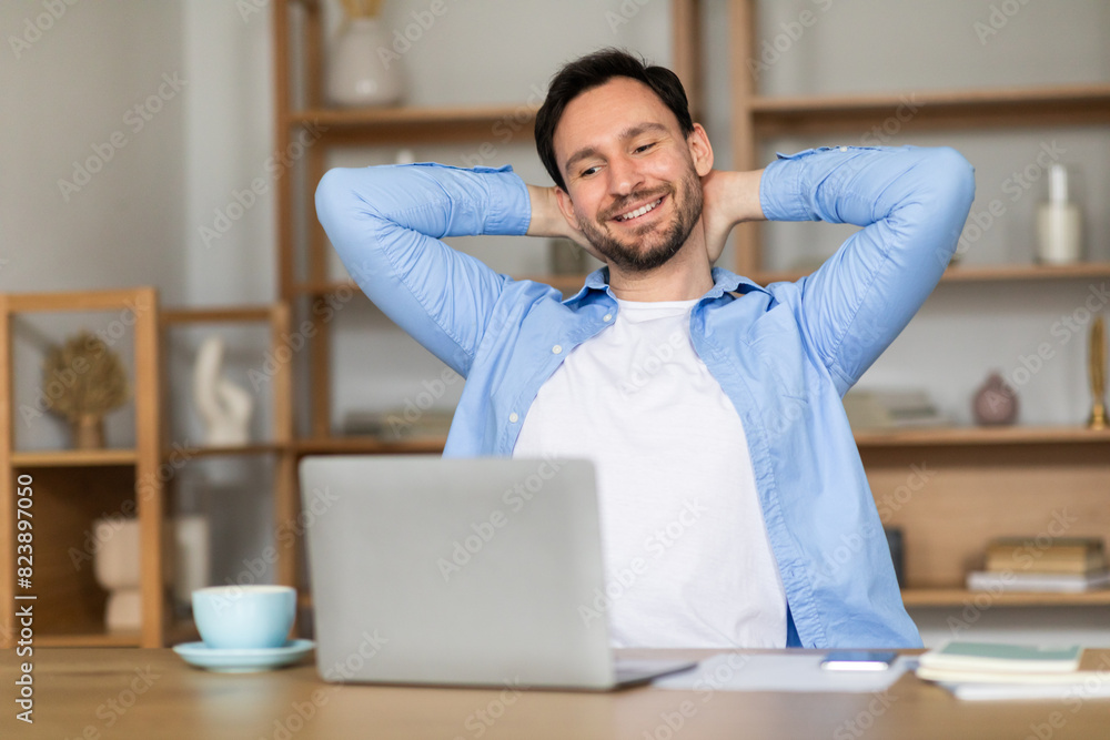 Obraz premium A content man leans back in his chair in a well-lit home office, his hands behind his head, expressing satisfaction and taking a break in front of his laptop, surrounded by notes and a cup of coffee.