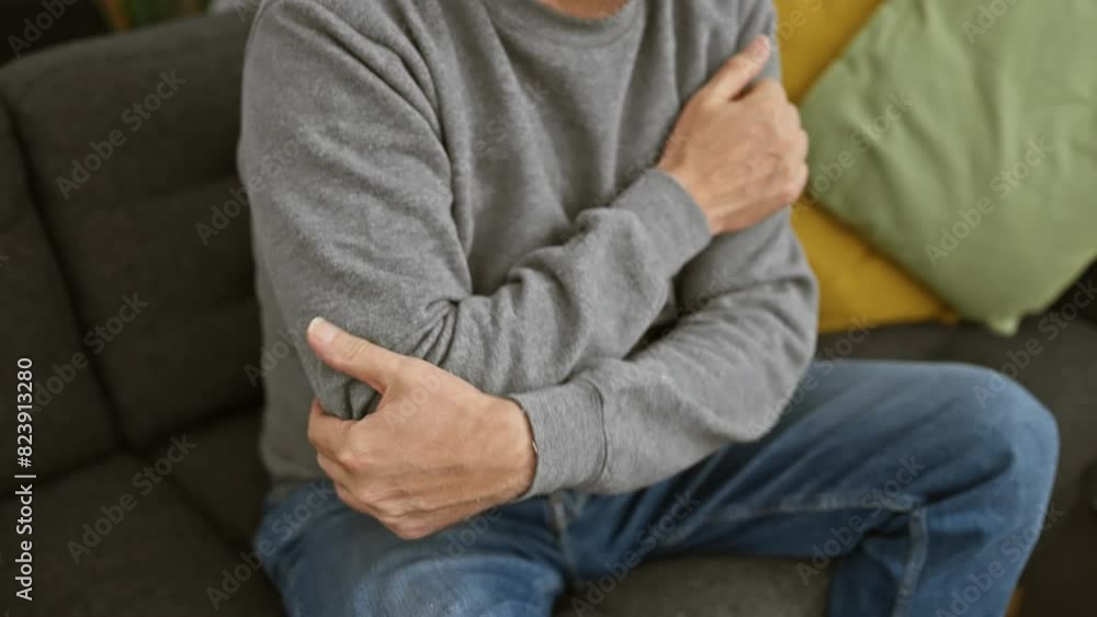 Middle-aged man in pain holding elbow inside a modern living room Middle-aged man in pain holding elbow inside a modern living room