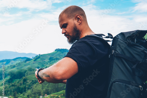 Pensive bearded tourist with backpack looking at smartwatch to tracking GPS walking in green locality of asia mountains.Hiker with rucksack managing tame on wristwatch during adventure trek photo