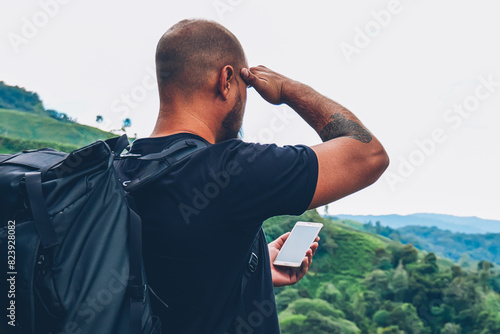 Back view of man hiker with travel backpack holding modern smartphone with navigator and looking in distance searching right direction in faraway places of beautiful green landscapes with high hills photo